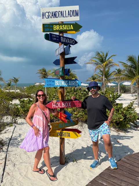 Leah in a pink and purple tie-dye sundress and Dave in flamingo shorts and a bucket hat stand smiling in front of the #GrandPalladium Costa Mujeres beach sign, with turquoise water and a bright sky in the background.